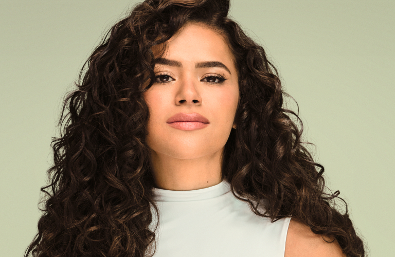 Foto com fundo verde claro. Mulher com cabelo cacheado e comprido, escuro, usando blusa branca sem mangas.